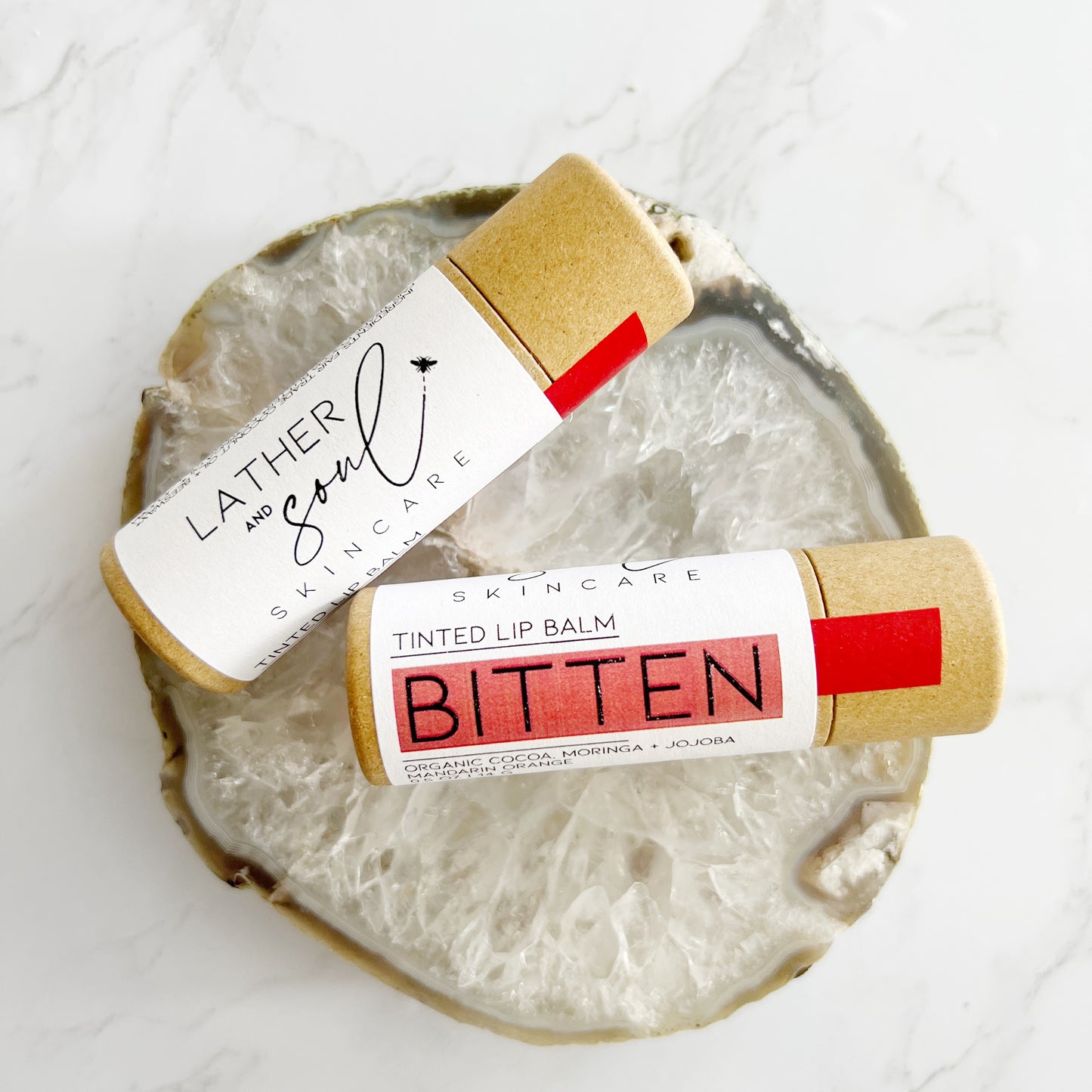 Natural and organic tinted lip balm, "Bitten," in a sheer crimson hue, packaged in eco-friendly paperboard tube.